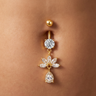 A gold-plated belly button ring with a round bezel-set clear gemstone and a dangling flower charm with pear-cut clear gemstones, shown worn on a person's abdomen.