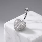 A silver-toned belly button ring with a heart-shaped pavé stone pendant and a small round stone at the end of the barbell, displayed on a white marble cube against a gray background.