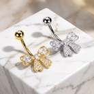 Two bow-shaped belly button rings displayed on a white marble cube. One ring is yellow gold with clear crystal pavé detailing, and the other is silver with matching clear crystal pavé detailing.