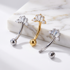Three curved barbell body jewelry piercings with clear faceted gemstone clusters at the top and ball ends, shown on a white marble block and white surface. Two are gold-toned and one is silver-toned, with a loose ball end placed in front.
