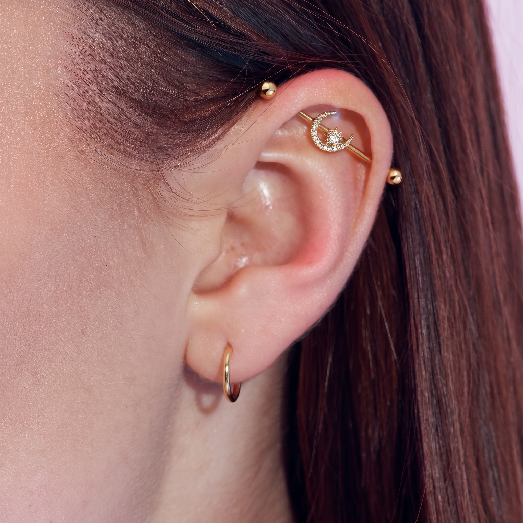 Close-up of a person's ear showcasing gold ear piercings, including a small hoop earring, a barbell stud, and a crescent moon cartilage piercing with a clear rhinestone center.