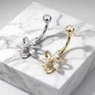Two curved barbell belly button piercings with flower-shaped rhinestone charms, one in silver and one in gold, displayed on a white marble block.