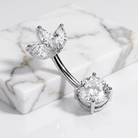 A silver-toned curved barbell belly button ring with marquise-cut clear gemstones on one end and a round clear gemstone on the other, displayed on a white marble block against a white background.