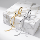 Two curved hoop earrings with bow and chain details, shown in gold and silver finishes on a white marble block against a light gray background.