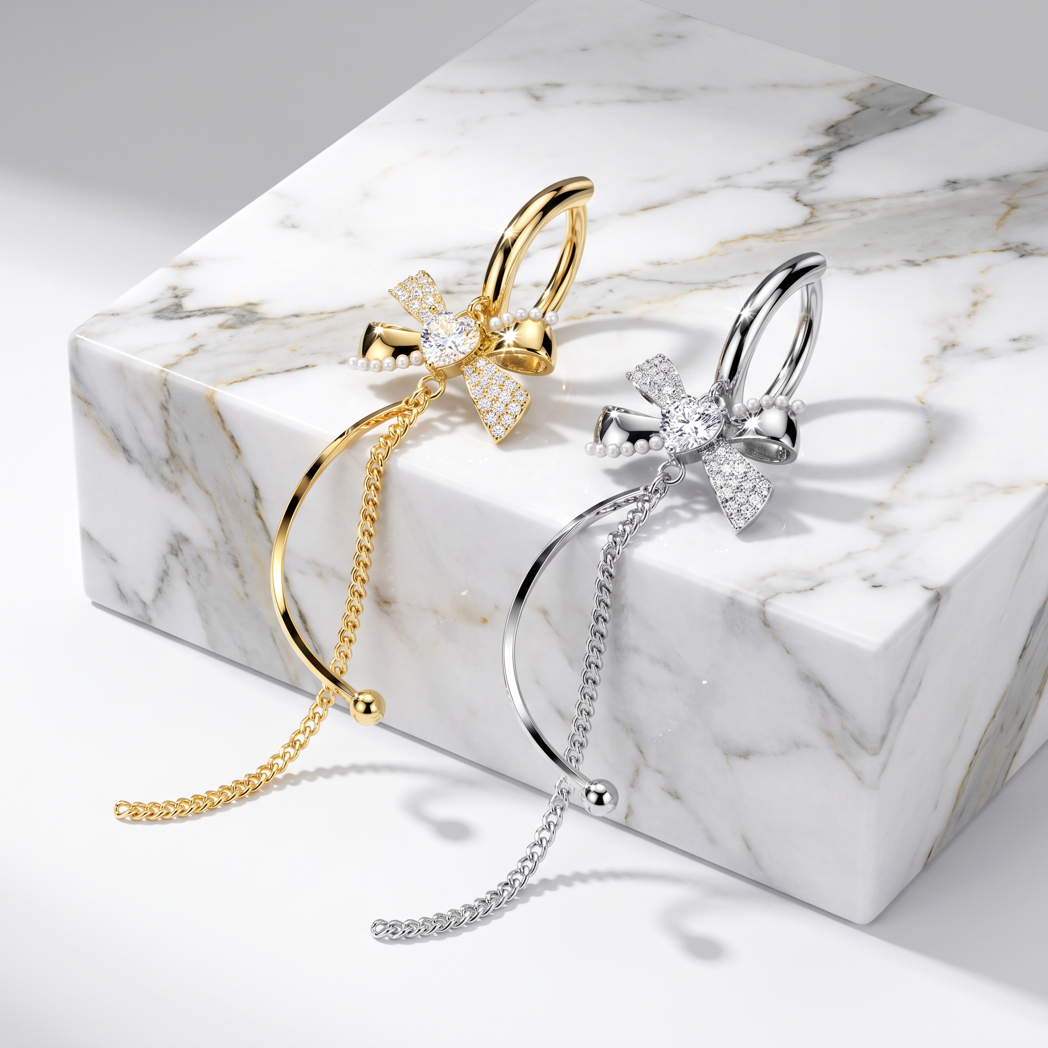 Two curved hoop earrings with bow and chain details, shown in gold and silver finishes on a white marble block against a light gray background.