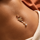 A close-up of a person's chest wearing a delicate gold belly button ring with a bow design and small clear gemstones, next to a matching gold stud earring.
