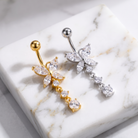 Two butterfly belly button rings displayed on a white marble slab. One ring is gold-plated with clear marquise and round crystals, and the other is silver-plated with matching clear crystals.