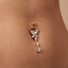 A close-up of a belly button piercing with a gold ball stud and a dangling gold belly button ring featuring marquise-cut clear crystals, shown on a model's skin.