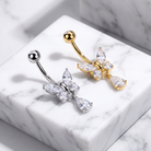 Two butterfly-shaped belly button rings displayed on a white marble block. One ring is silver-toned with clear marquise and round stones, and the other is gold-toned with matching clear stones.