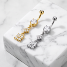 Two belly button rings with dangling floral and chain details, one in yellow gold and one in silver, displayed on a white marble block.