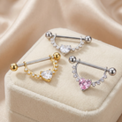 Three curved barbell piercings with heart-shaped clear and pink stones, displayed on a cream velvet jewelry box against a soft beige satin background.