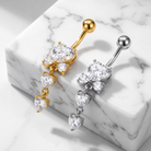 Two belly button rings with dangling heart-shaped clear gemstones, shown in gold and silver metal finishes, displayed on a white marble block.