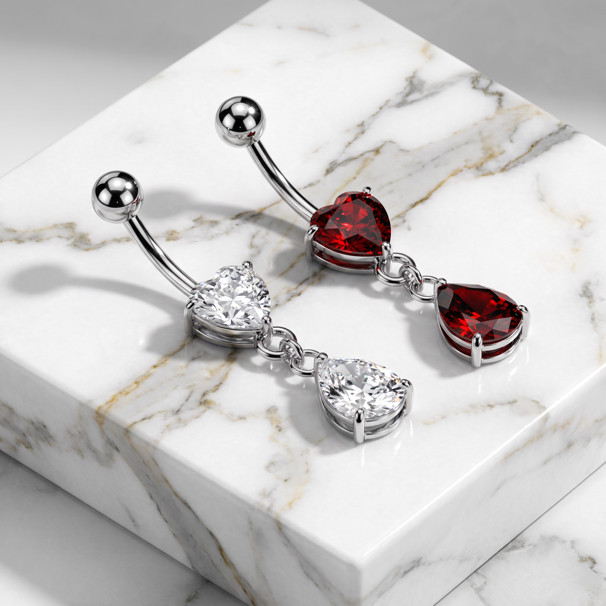 A pair of silver-toned belly button rings with heart-shaped red gemstones and teardrop-shaped clear gemstones, displayed on a white marble block.