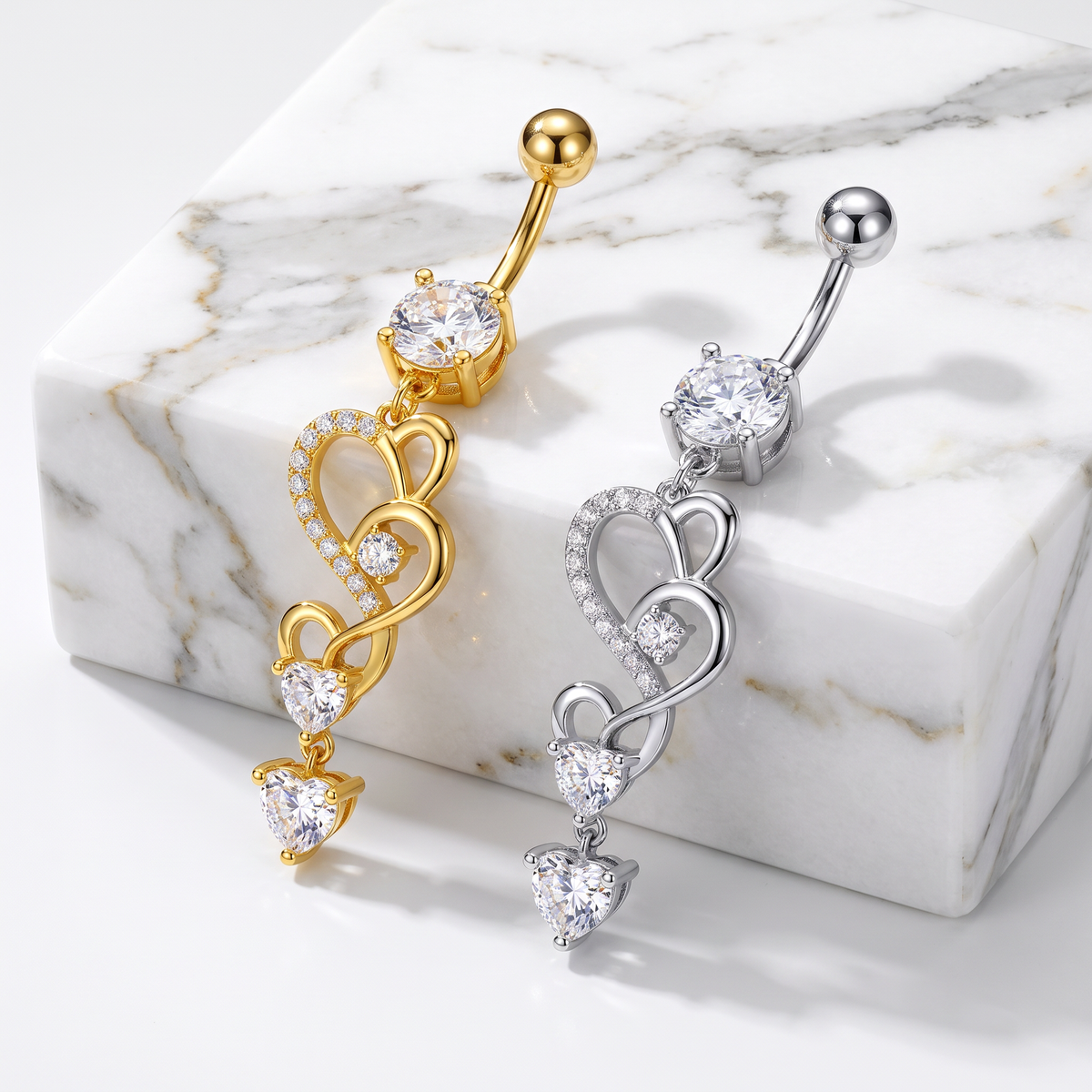 Two heart-themed belly button rings, one in gold and one in silver, both featuring a large round clear stone and three cascading heart designs with small clear stones, displayed on a white marble block.