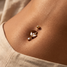 A close-up of a person's navel wearing a gold leaf-shaped belly button ring with clear rhinestones and a small gold bead stud nearby, against warm skin tones and a beige garment.