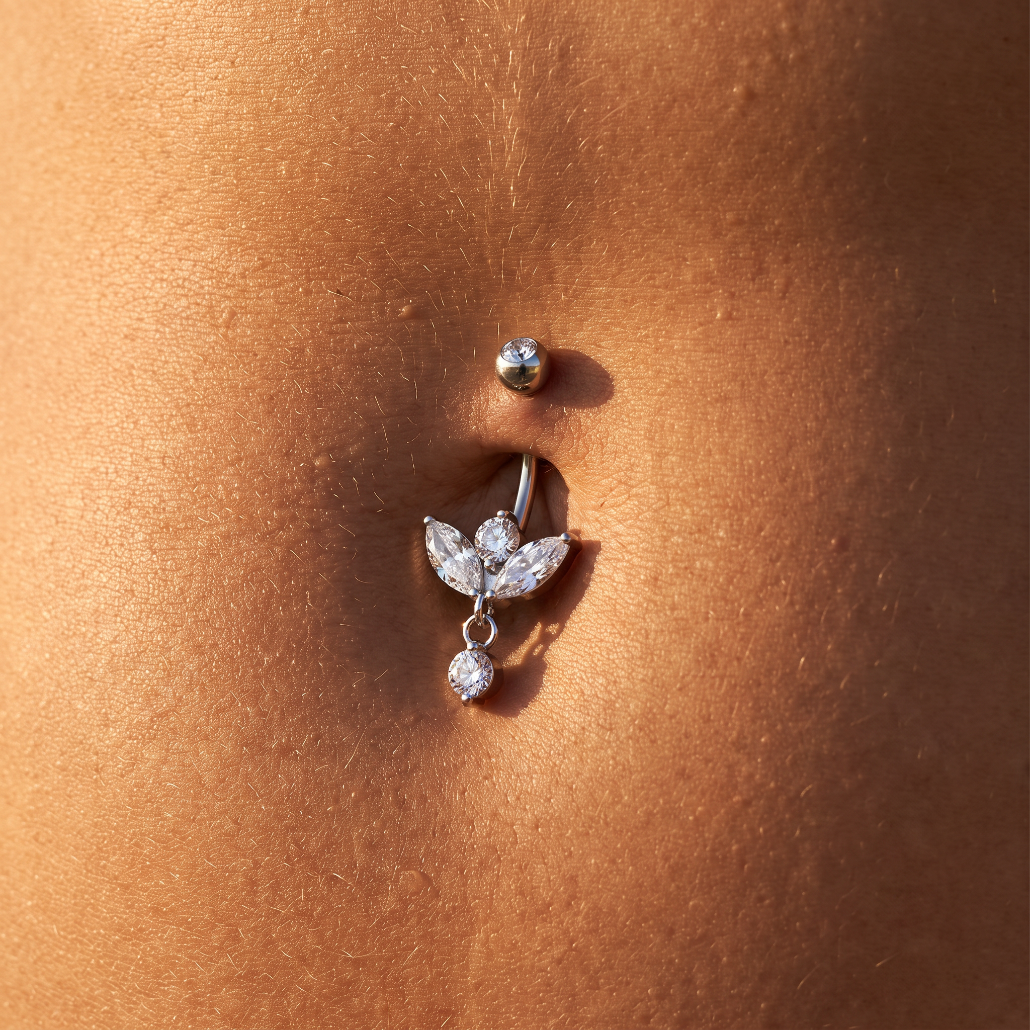 A close-up of a silver belly button ring with clear rhinestones, worn on a person's navel against warm skin tones.