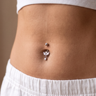 A close-up of a person's midsection showing a silver belly button ring with a small top stud and a dangling butterfly-shaped charm, worn with a white ribbed crop top and white shorts.