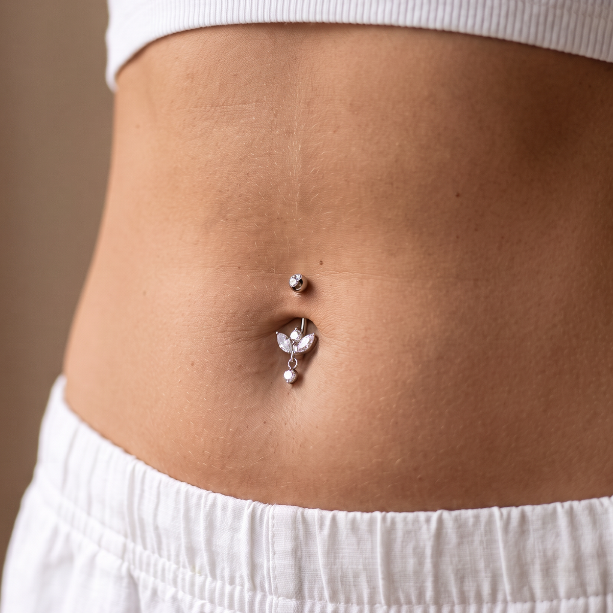 A close-up of a person's midsection showing a silver belly button ring with a small top stud and a dangling butterfly-shaped charm, worn with a white ribbed crop top and white shorts.
