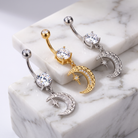 Three belly button rings with crescent moon and star charms are displayed on a white marble block. Each ring features a silver or gold curved barbell, a round clear crystal, and a dangling crescent moon charm embellished with small clear crystals and a star.