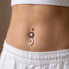 A close-up of a person's midsection showing a silver belly button ring with a dangling crescent moon charm and a small clear rhinestone accent, worn with a white ribbed crop top and white shorts.