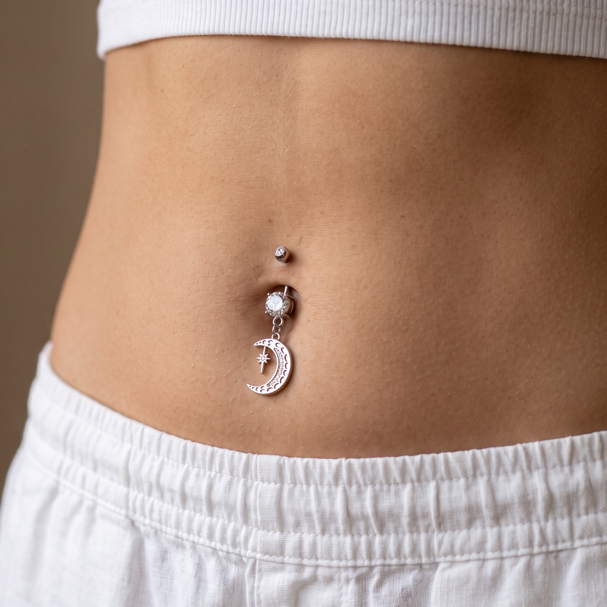 A close-up of a person's midsection showing a silver belly button ring with a dangling crescent moon charm and a small clear rhinestone accent, worn with a white ribbed crop top and white shorts.