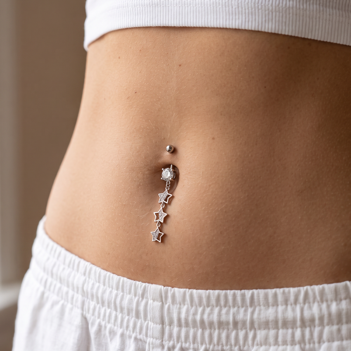A close-up of a person's midsection wearing a silver belly button ring with a dangling star charm, styled with a white ribbed crop top and white sweatpants.