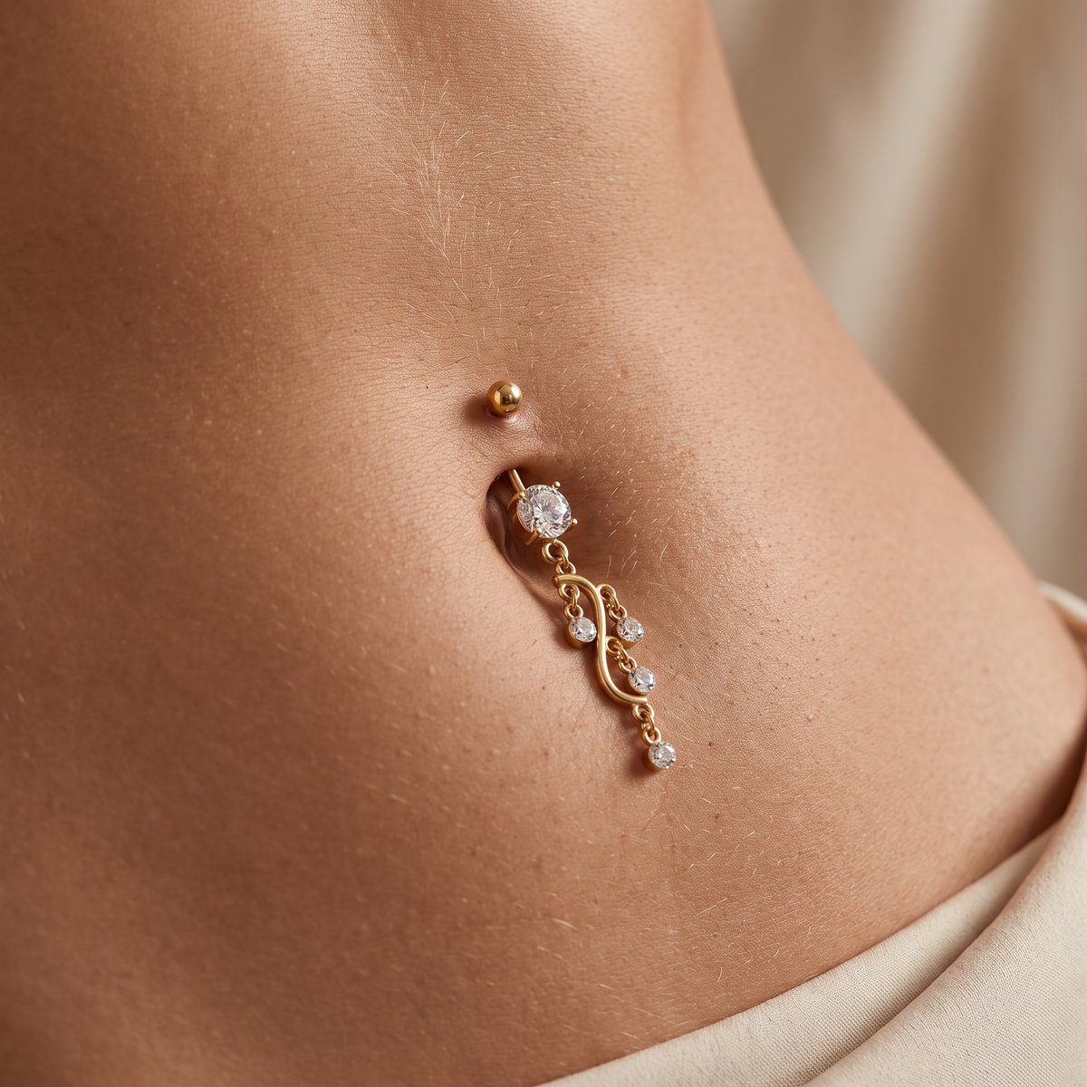A close-up of a person's lower torso showing a gold belly button ring with a dangling scroll design and clear gemstones, along with a matching gold belly button stud.