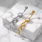 Two curved belly button rings with ornate dangling chains and clear gemstones, shown on a white marble block. One ring is silver-toned and the other is gold-toned.