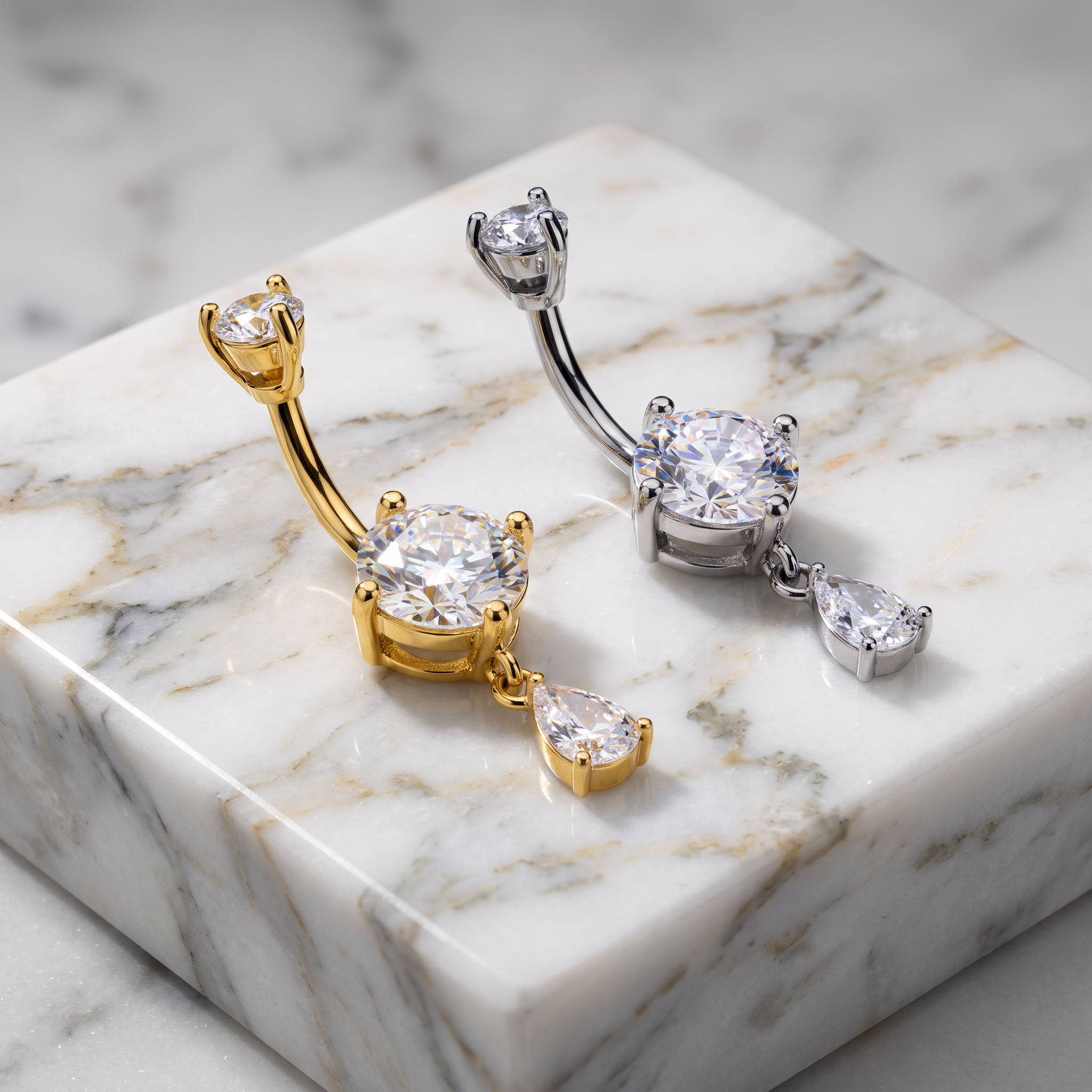 Two curved barbell belly button piercings with large round clear gemstones and teardrop dangling stones, shown in yellow gold and silver metal on a white marble block.