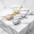 Two teardrop-shaped belly button rings with halo-set clear stones, one in yellow gold and one in silver, displayed on a white marble block.