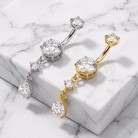 Two curved barbell belly button piercings with large round and teardrop-cut clear gemstones, shown on a white marble block. One is silver-toned and the other is yellow gold-toned.