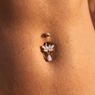 A close-up of a belly button piercing with a gold flower-shaped stud set with clear crystals and a dangling teardrop crystal, shown on skin.