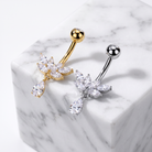 Two flower-shaped belly button rings with clear crystal petals, one in gold-tone metal and one in silver-tone metal, displayed on a white marble cube.