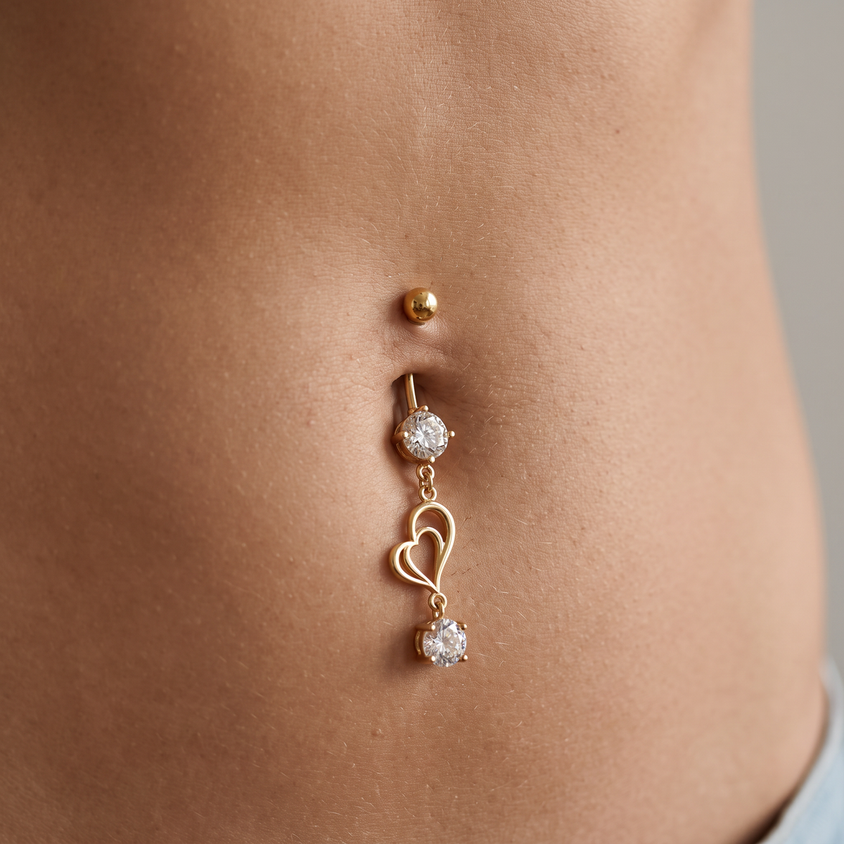 A gold belly button ring with a small ball stud, a round crystal-set post, a dangling heart outline, and a round crystal accent, shown worn on a person's abdomen.
