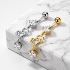 Two curved barbell body jewelry piercings with double heart charms and clear gemstones, shown on a white marble block. One is silver-toned and one is gold-toned.