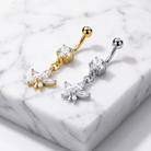 Two belly button ring piercings displayed on a white marble block. One is gold-toned with a clear crystal and dangling flower charm, and the other is silver-toned with a clear crystal and matching flower charm.