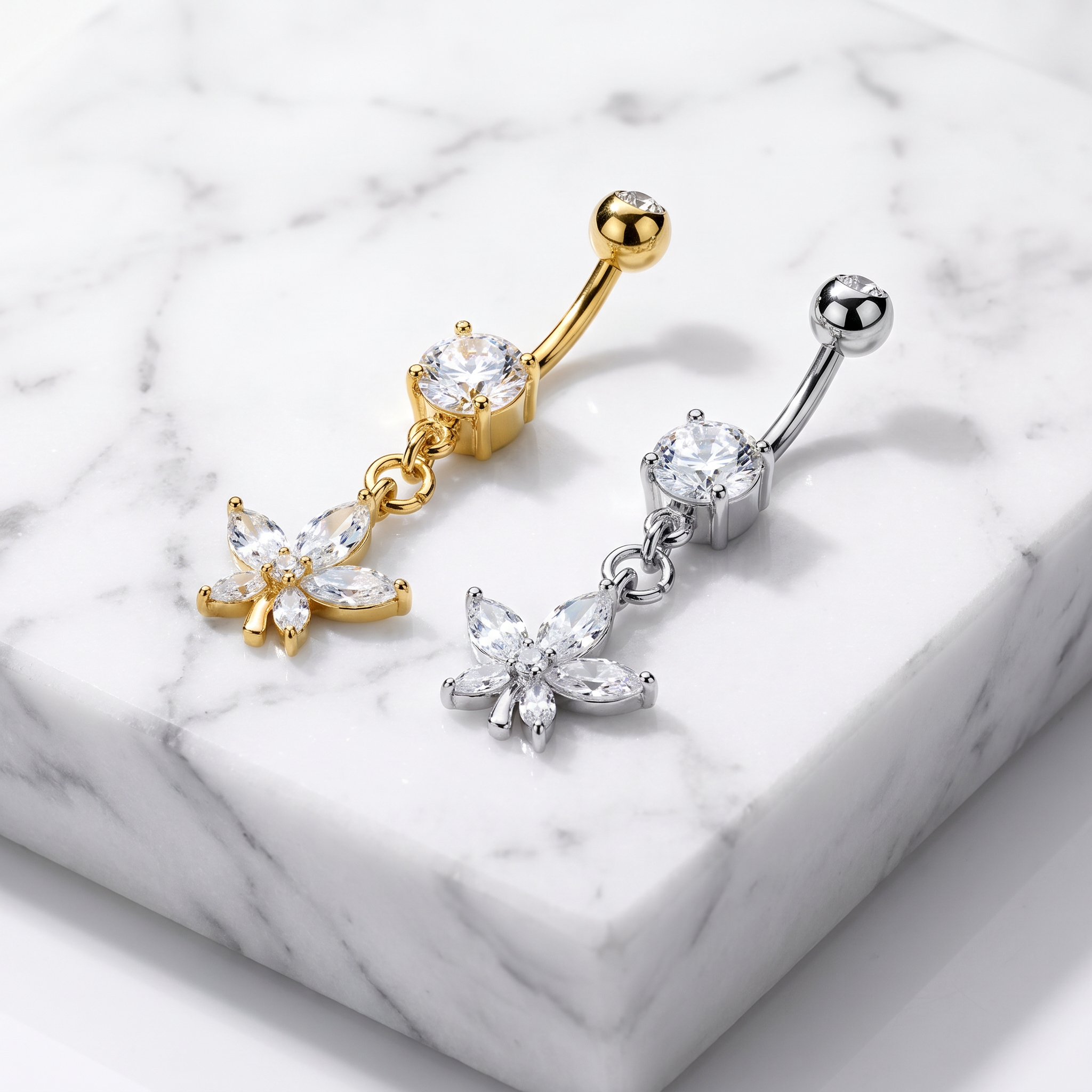 Two belly button ring piercings displayed on a white marble block. One is gold-toned with a clear crystal and dangling flower charm, and the other is silver-toned with a clear crystal and matching flower charm.