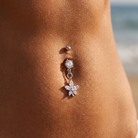A silver belly button ring with a dangling flower charm and clear rhinestones, shown worn on a person's navel with a blurred beach background.