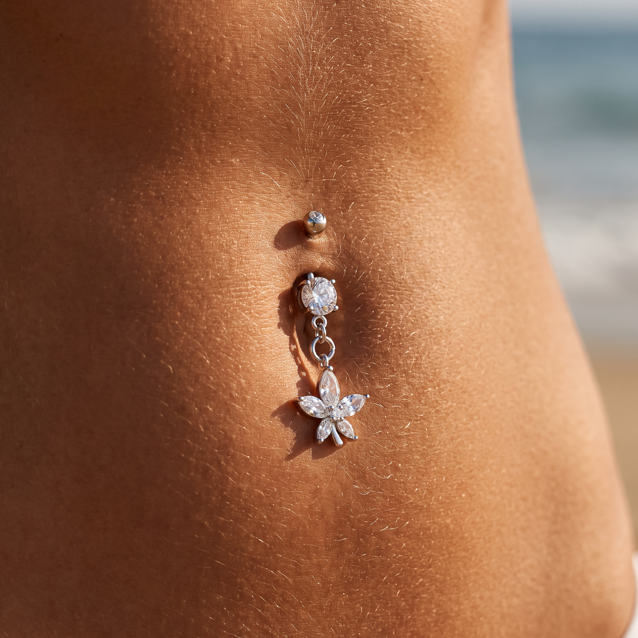A silver belly button ring with a dangling flower charm and clear rhinestones, shown worn on a person's navel with a blurred beach background.