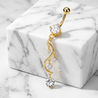 A gold belly button ring with a dangling wavy chain and clear cubic zirconia stones, displayed on a white marble block over a marble surface.