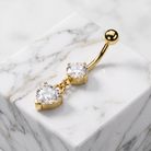 Gold-plated belly button ring with two clear heart-shaped gemstones, displayed on a white marble block.