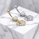 Two curved barbell belly button piercings with heart-shaped crystal clusters and dangling heart charms, shown in gold and silver on a white marble pedestal.
