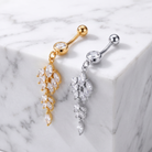 Two leaf-shaped belly button rings with marquise-cut clear crystals, one in gold-tone metal and one in silver-tone metal, displayed on a white marble block.