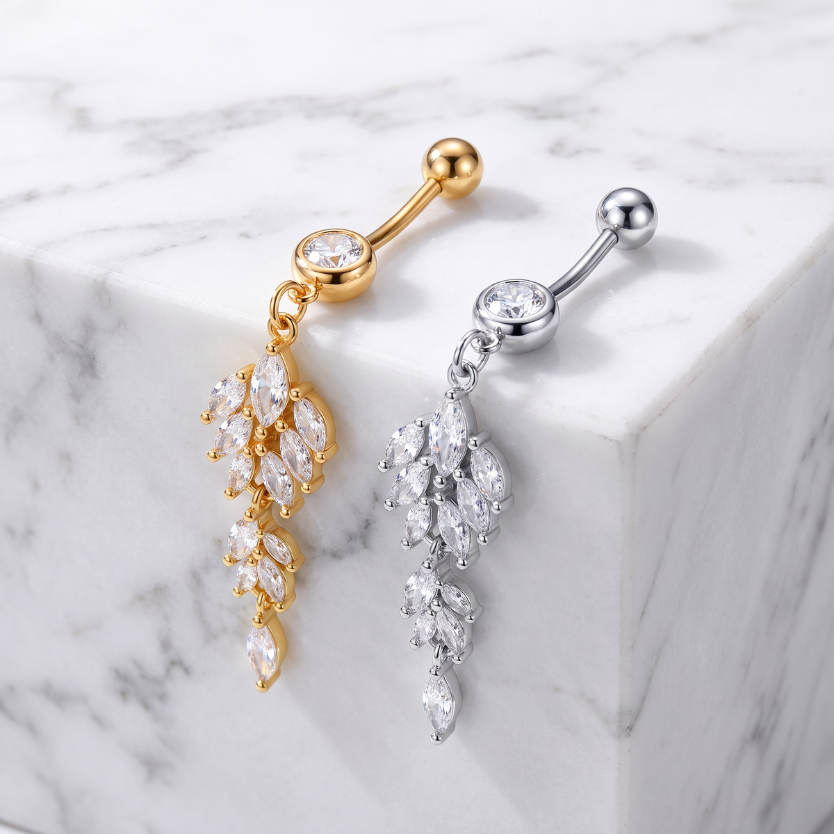 Two leaf-shaped belly button rings with marquise-cut clear crystals, one in gold-tone metal and one in silver-tone metal, displayed on a white marble block.