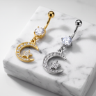 Two belly button rings with crescent moon and star charms are displayed on a white marble block. One ring is gold-toned with a clear cubic zirconia stone and a gold crescent moon set with small clear stones, while the other is silver-toned with a matching silver crescent moon and stone.