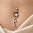 A silver-toned belly button ring with a dangling moon and star charm, adorned with clear rhinestones, shown worn on a model's navel with a beige satin fabric in the foreground.