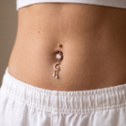 A close-up of a person's midsection wearing a rose gold belly button ring with a dangling crescent moon and teardrop charm, along with a small gold ball stud.