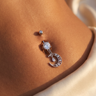 A close-up of a person's navel wearing a silver-toned belly button ring with a clear round gemstone and a dangling crescent moon charm studded with small clear stones.