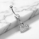 A silver-toned belly button ring with a curved barbell, a round clear gemstone, and a dangling crown charm adorned with small clear stones, displayed on a white marble surface.