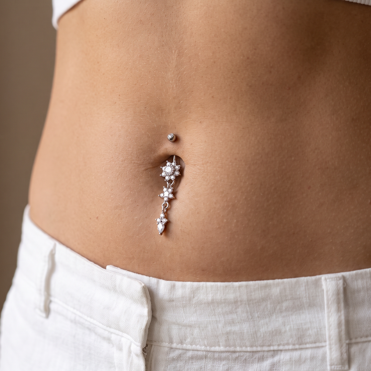 A close-up of a person's lower torso wearing a silver belly button ring with clear rhinestones, styled with a white ribbed crop top and white linen pants.